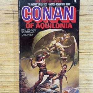 May include: Vintage paperback book cover for "Conan of Aquilonia." The cover features a muscular warrior wielding a sword, a young man with a spear, and a winged creature. The title is in large red and purple letters. The book is by L. Sprague de Camp and Lin Carter.
