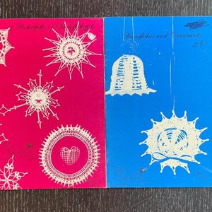 Op de afbeelding: Twee vintage boekjes met witte kanten sneeuwvlok- en ornamentontwerpen. Eén boekje is roze met de tekst "2 Pocketfulls of Snowflakes". De andere is blauw met de tekst "Snowflakes and Ornaments".