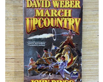 Op de afbeelding: Een paperback boek getiteld "March Upcountry" van David Weber en John Ringo. De omslag toont een sciencefiction-illustratie van soldaten met wapens die op dinosaurusachtige wezens rijden. Het boek heeft een donkere achtergrond met de tekst "Superb Storytelling!" en het logo van de uitgever.
