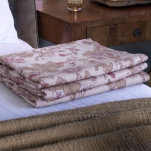 May include: A folded fabric textile featuring a light cream background with a repeating floral pattern in shades of muted pink, mauve, and soft brown. The fabric is neatly stacked in layers, displaying a botanical print with leaves and blossoms.