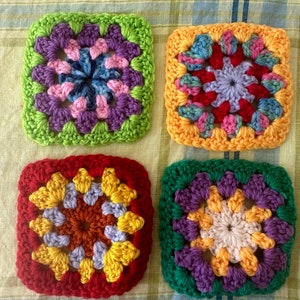 May include: Four crocheted squares in different colors, each with a unique flower design in the center. The squares are green, orange, red, and purple.