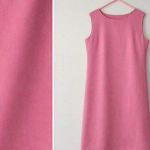 May include: A sleeveless, knee-length pink dress hangs on a pink hanger. The dress has a simple, straight silhouette with a round neckline. The fabric appears to be a solid colour, with a subtle texture. The dress is displayed against a white background.