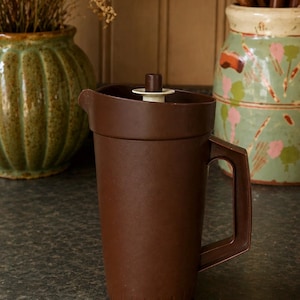 May include: A dark brown plastic jug featuring a matching lid with a small, central white and brown plunger mechanism. The jug has a sturdy, angular handle on the side and a simple pouring spout at the top rim, with vertical ridges at the base.