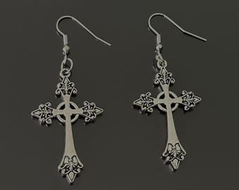 May include: A pair of silver-colored cross earrings. Each earring features a detailed cross design with ornate embellishments. The earrings hang from simple fish hook ear wires, suitable for pierced ears. The background is a dark, neutral tone.