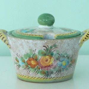 May include: A white ceramic sugar bowl with a green lid and floral design. The bowl has two yellow and brown handles and a small spout on the lid.