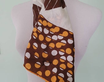 Può includere: Un foulard di seta bianco con un motivo geometrico marrone, arancione e bianco. Il foulard è legato intorno al collo di un manichino bianco.