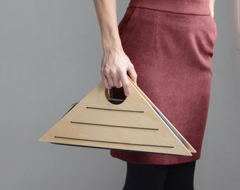 May include: A triangular handbag with a handle is held. The bag is light-colored with a geometric design. The person is wearing a black vest top and a burgundy skirt. The bag's design features horizontal lines.