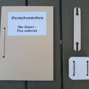 Könnte beinhalten: Ein braunes Heft mit dem Titel "Brettchenweben" und dem Text "Alte Kunst - Neu entdeckt". Das Heft ist mit einer Schnur gebunden und hat zwei schwarze Punkte auf dem Umschlag. Neben dem Heft liegen zwei Holzstücke.