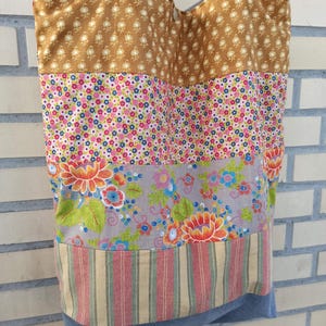 May include: A patchwork tote bag with a variety of floral and patterned fabrics. The top section is a tan fabric with a small white floral pattern. Below are sections of pink floral, gray floral, and striped fabric. The bottom is a blue denim.