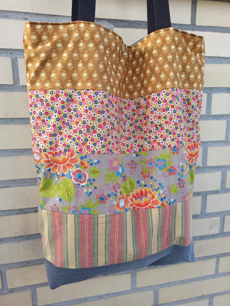 May include: A patchwork tote bag with a variety of floral and patterned fabrics. The top section is a tan fabric with a small white floral pattern. Below are sections of pink floral, gray floral, and striped fabric. The bottom is a blue denim.