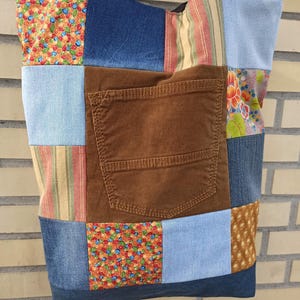 Pu&ograve; includere: Una borsa tote patchwork con una variet&agrave; di tessuti, tra cui denim, velluto a coste e cotone fantasia. La borsa presenta un'ampia tasca in velluto a coste marrone e una tracolla nera. Il design patchwork incorpora tessuti blu, marroni e fantasia.