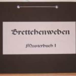Könnte beinhalten: Ein schwarzer Ordner mit einem weißen rechteckigen Etikett. Das Etikett trägt den Text "Brettchenweben" in gotischer Schrift über "Musterbuch 1". Oben befindet sich eine silberne Öse.