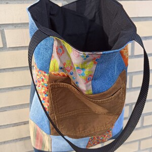 Pu&ograve; includere: Una borsa tote patchwork con una tasca in velluto a coste marrone e cinghie nere. L'esterno della borsa presenta un mix di denim e tessuti fantasia in vari colori, tra cui blu, arancione e stampe floreali. L'interno &egrave; nero.
