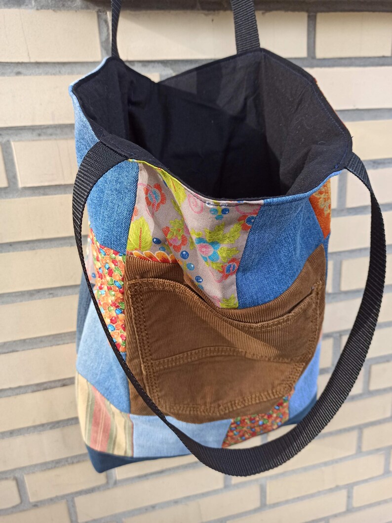 Pu&ograve; includere: Una borsa tote patchwork con una tasca in velluto a coste marrone e cinghie nere. L'esterno della borsa presenta un mix di denim e tessuti fantasia in vari colori, tra cui blu, arancione e stampe floreali. L'interno &egrave; nero.