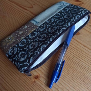 Może przedstawiać: Etui na oł&oacute;wki z brązowej i beżowej tkaniny patchworkowej z zamkiem błyskawicznym. Z etui wystaje niebieski długopis.