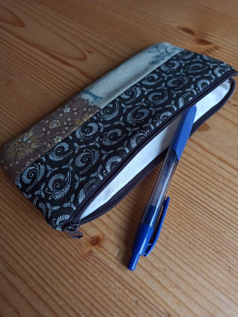 Może przedstawiać: Etui na oł&oacute;wki z brązowej i beżowej tkaniny patchworkowej z zamkiem błyskawicznym. Z etui wystaje niebieski długopis.