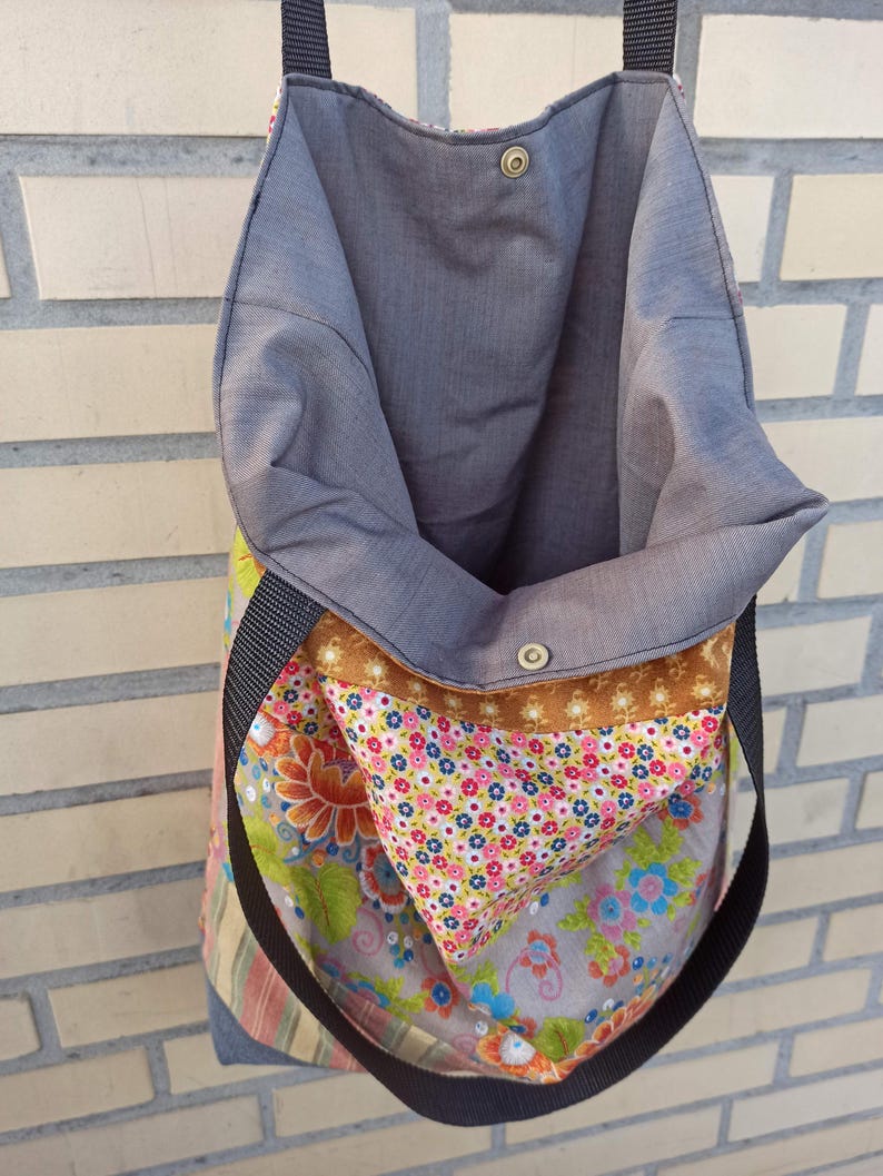 May include: Open tote bag with a floral pattern and a gray interior. The bag has a black strap and a brown accent. The exterior features a mix of floral and striped fabrics, with a snap closure at the top.