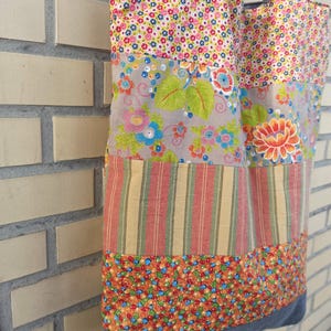 May include: A patchwork tote bag with a variety of floral and striped patterns in shades of pink, blue, orange, and green. The bag has black handles and a gray denim base. The bag is made of fabric.