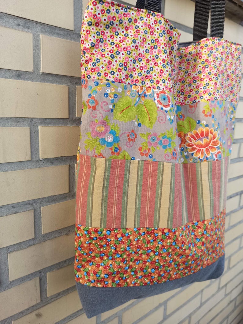 May include: A patchwork tote bag with a variety of floral and striped patterns in shades of pink, blue, orange, and green. The bag has black handles and a gray denim base. The bag is made of fabric.