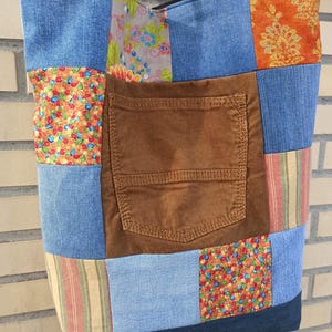 Könnte beinhalten: Eine Patchwork-Tragetasche mit verschiedenen Stoffen, darunter Jeans, Cord und gemusterte Baumwolle. Die Tasche hat eine braune Cordtasche und einen schwarzen Riemen. Das Design beinhaltet blaue, orange, rote und grüne Farben.