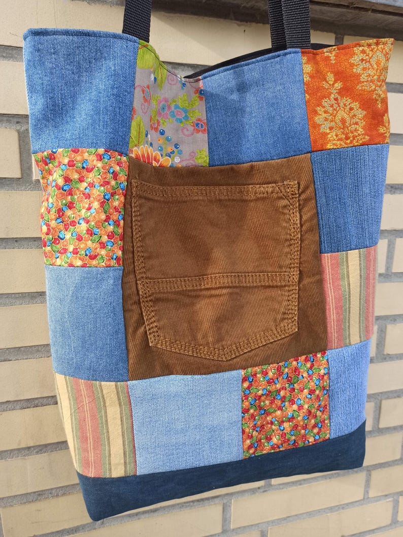 Pu&ograve; includere: Una borsa tote patchwork con una variet&agrave; di tessuti, tra cui denim, velluto a coste e cotone fantasia. La borsa presenta una tasca in velluto a coste marrone e una tracolla nera. Il design incorpora colori blu, arancione, rosso e verde.