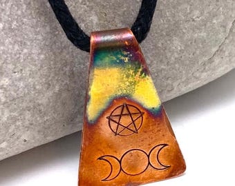 May include: A trapezoidal copper pendant suspended from a black cord. The metal features a heat-treated finish with gradients of yellow, green, and orange. A pentagram is etched above a triple moon symbol on the front surface of the copper piece.