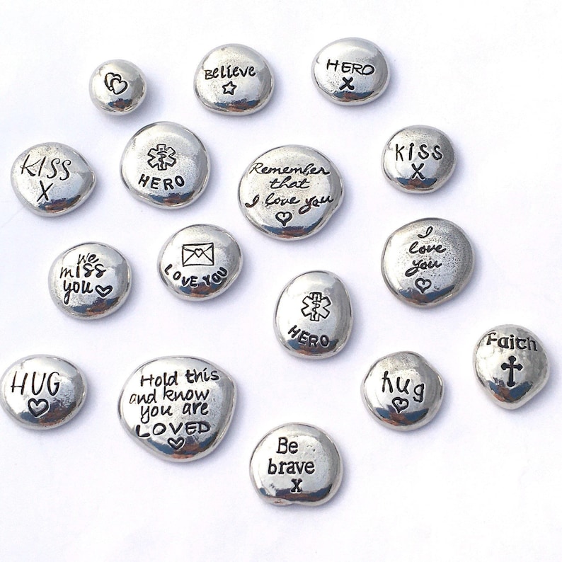 A selection of shiny pewter pebbles, each one hand stamped with different sayings and symbols, like remember that I love you, hold this and know you are loved, hug, we miss you, faith with crucifix symbol. Pebbles are displayed on a white background.