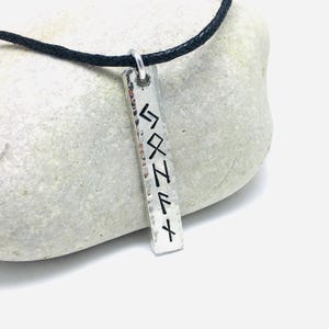 Pewter bar with square ends and hammered edges hangs from 2mm black cotton cord. It is resting against a pale grey rock. Bar is 1cm wide and 6mm viking runes have been stamped vertically to form a name.