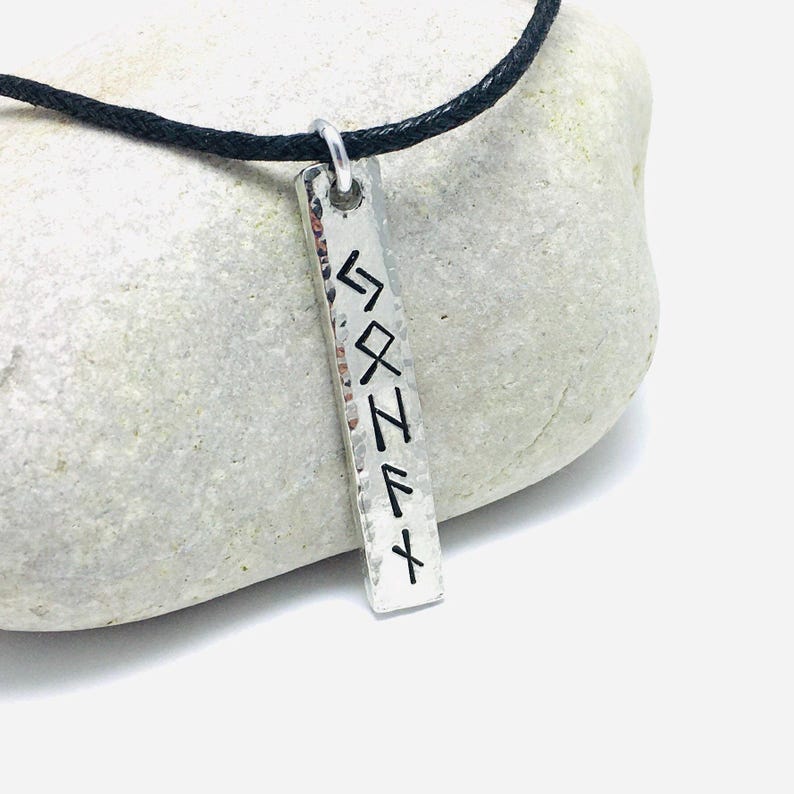 Pewter bar with square ends and hammered edges hangs from 2mm black cotton cord. It is resting against a pale grey rock. Bar is 1cm wide and 6mm viking runes have been stamped vertically to form a name.