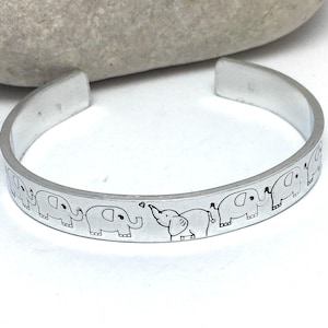 Puede incluir: Una pulsera de plata con un patrón repetido de elefantes grabados. La pulsera abierta está hecha de un material metálico brillante. Los elefantes están representados en un estilo de dibujos animados simple, con sus trompas levantadas.