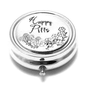 Puede incluir: Un pastillero redondo de color plateado con la inscripción "Happy Pills". La tapa presenta diseños florales y de hojas. El pastillero tiene un pequeño botón plateado para abrirlo.