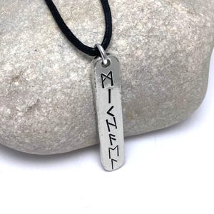 Pewter bar with round ends and hammered edges hangs from 2mm black cotton cord. It is resting against a pale grey rock. Bar is 1cm wide and 6mm viking runes have been stamped vertically to form a name.