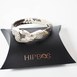 Peut inclure: Un bracelet en peau de serpent avec un fermoir argenté, posé sur une boîte noire. Le bracelet présente un motif d'écailles marron, grises et blanches. La boîte porte l'inscription "HIPBOS".