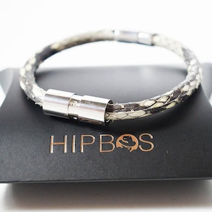 Op de afbeelding: Een zwarte leren armband met een zilveren magneetsluiting. De armband ligt op een zwarte doos met het woord "HIPBOS" in goud gedrukt.