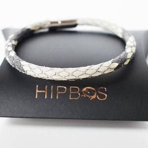 May include: A snake skin bracelet with a magnetic clasp sits on a black triangular box. The bracelet has a pattern of white, grey, and black scales. The box has the brand name "HIPBOS" in copper-coloured lettering.