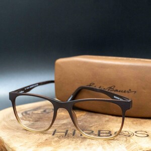 Könnte beinhalten: Braune rechteckige Brille mit Farbverlaufgläsern, auf einer Holzoberfläche. Im Hintergrund ein braunes Lederetui mit dem Eddie Bauer-Logo. Die Brille hat ein modernes, minimalistisches Design.