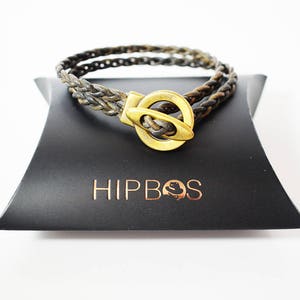 以下が含まれることがあります： 金色の留め具が付いた編み込みの革製のブレスレット。ブレスレットは、「HIPBOS」という文字が印刷された黒い箱の上に置かれています。