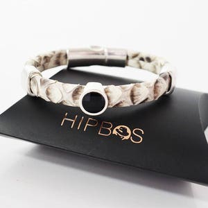 Op de afbeelding: Een wit leren armband met een zwarte onyx steen en een zilveren magneetsluiting. De armband ligt op een zwarte achtergrond met het woord "HIPBOS" in goud gedrukt.