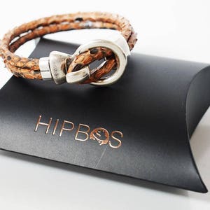 Può includere: Un braccialetto in pelle marrone con una chiusura argentata. Il braccialetto è su un cuscino nero con la scritta "HIPBOS" stampata su di esso.