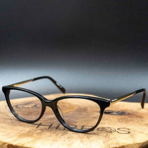 Puede incluir: Gafas negras de ojo de gato con detalles dorados en las patillas. Las monturas se presentan sobre una superficie de madera con un fondo oscuro. Las gafas tienen un diseño moderno y elegante.