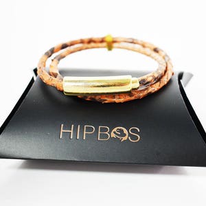 Op de afbeelding: Een bruin leren armband met een gouden sluiting. De armband ligt op een zwarte achtergrond met het woord "HIPBOS" in goud gedrukt.
