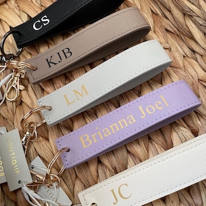 May include: Several personalised keyrings in black, beige, white, and lilac. Each keyring has gold-coloured metal hardware and is embossed with initials or a name. The keyrings are displayed on a woven, natural-toned surface.