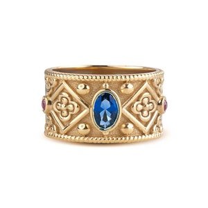 May include: A wide gold band ring featuring a central oval blue gemstone, flanked by four-petaled floral motifs within diamond-shaped frames, accented with small round red stones on the sides and a beaded texture along the top and bottom edges.