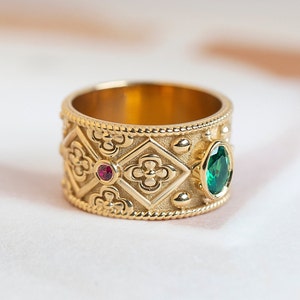 May include: A wide gold band ring featuring a textured surface with repeating diamond-shaped patterns containing embossed quatrefoil motifs. The ring is accented with a small round red gemstone and a larger oval green gemstone set in gold bezels.