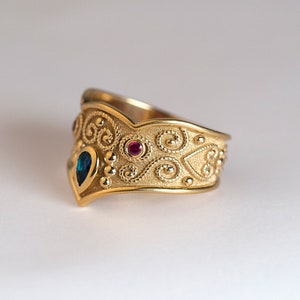 May include: A gold ring featuring a textured surface with intricate swirling filigree patterns, small raised metal beads, a central pear-shaped blue gemstone, and a round red gemstone set into the band with a pointed front edge design.