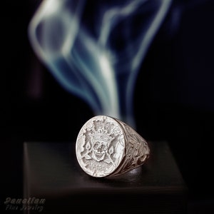 Puede incluir: Un anillo de sello de plata con una cara circular que muestra un cr&aacute;neo grabado con una corona, flanqueado por motivos florales. La banda presenta intrincados patrones de volutas en los laterales. El texto Danelian Fine Jewelry aparece en la esquina.