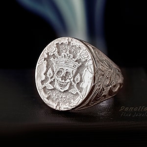 May include: A silver signet ring with a detailed design of a skull wearing a crown and surrounded by roses. The ring band is also intricately engraved.