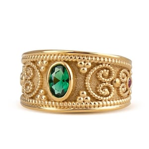 May include: A gold ring with an intricate design featuring a green oval gemstone and two smaller red gemstones. The ring has a textured surface and a wide band.