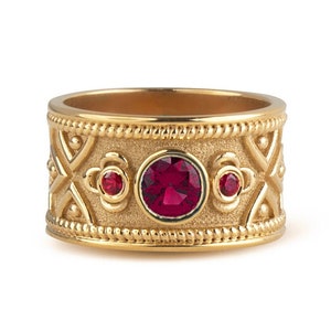 May include: A gold ring with a red gemstone and intricate detailing. The ring has a textured surface and a band with a repeating pattern.