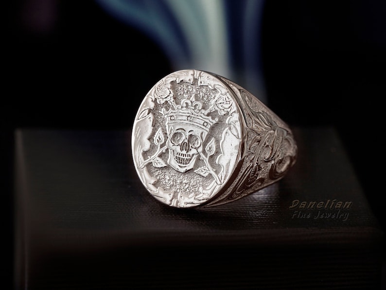 May include: A silver signet ring with a detailed design of a skull wearing a crown and surrounded by roses. The ring band is also intricately engraved.